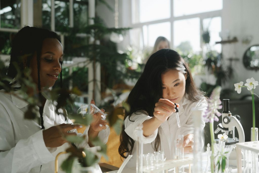 Two women in lab coats conducting an experiment in a sunlit indoor laboratory setting.